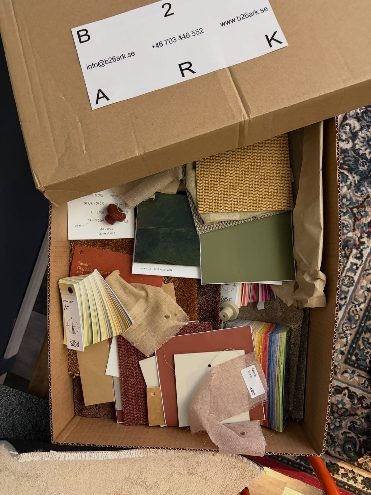 Öppen kartong märkt B26 ARK full av materialprover: tyger, kakel, mattbitar och färgkartor